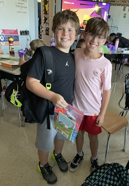 Two young boys in a classroom, one holding a plastic bag and the other wearing glasses. Tables, chairs, and a monitor are in the background.