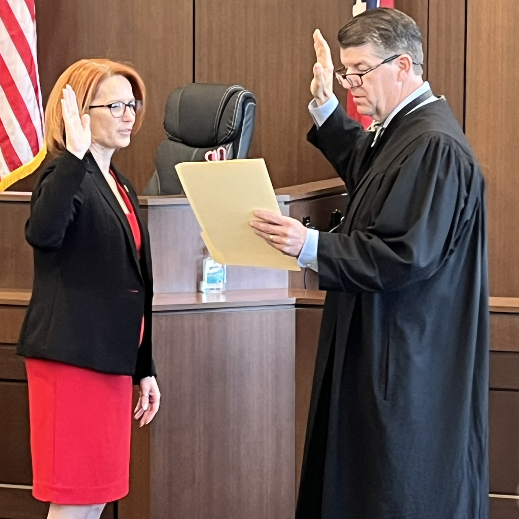 A woman in a red skirt and black blazer takes an oath with a raised hand. A man in a black robe stands next to her.