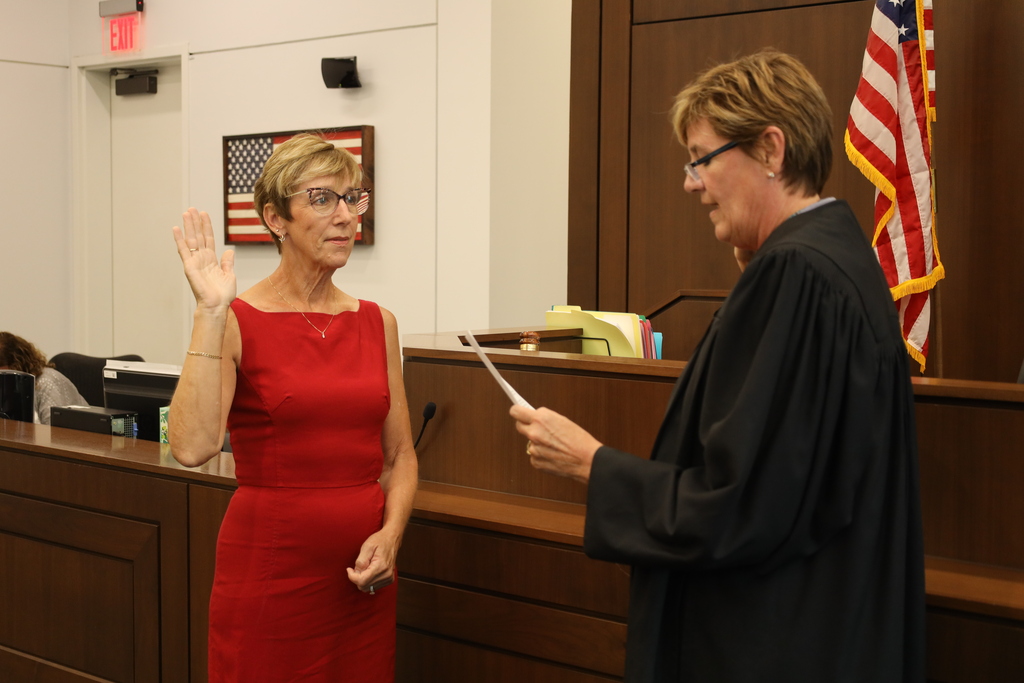 Two women stand in a courtroom; one in red raising her hand, the other in black holding a paper. Behind them is a judge's bench and a flag.