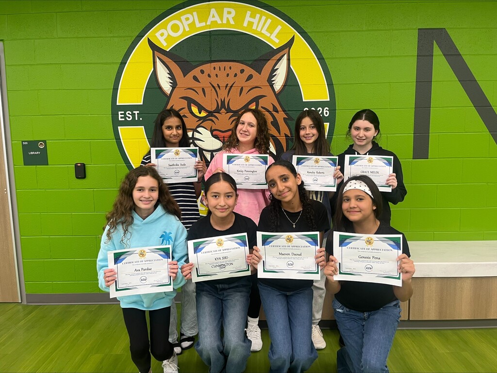 Nine children pose for a photo holding certificates in front of a green wall with a school logo.