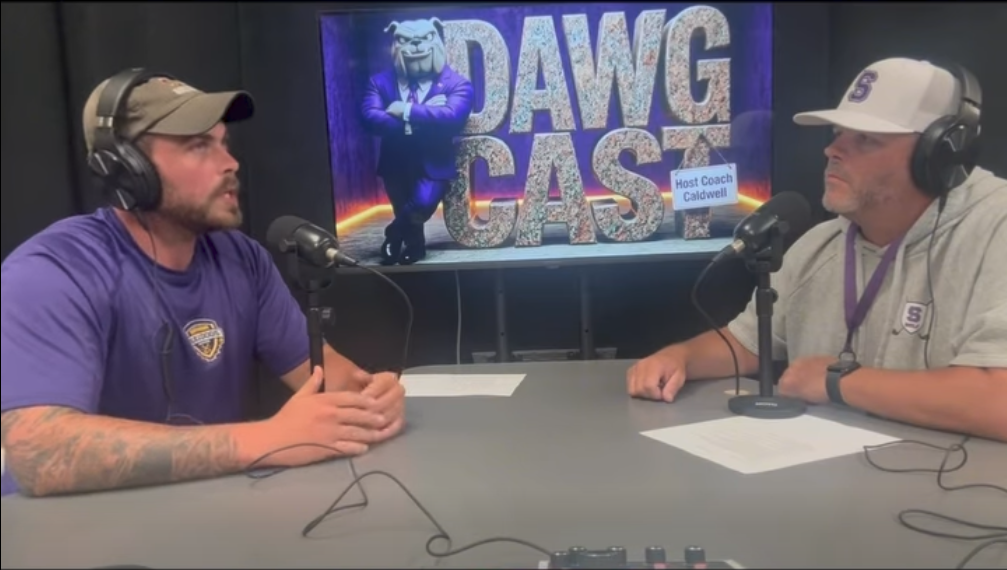 Two men seated at a table with microphones, one in a purple shirt, the other in a gray shirt.