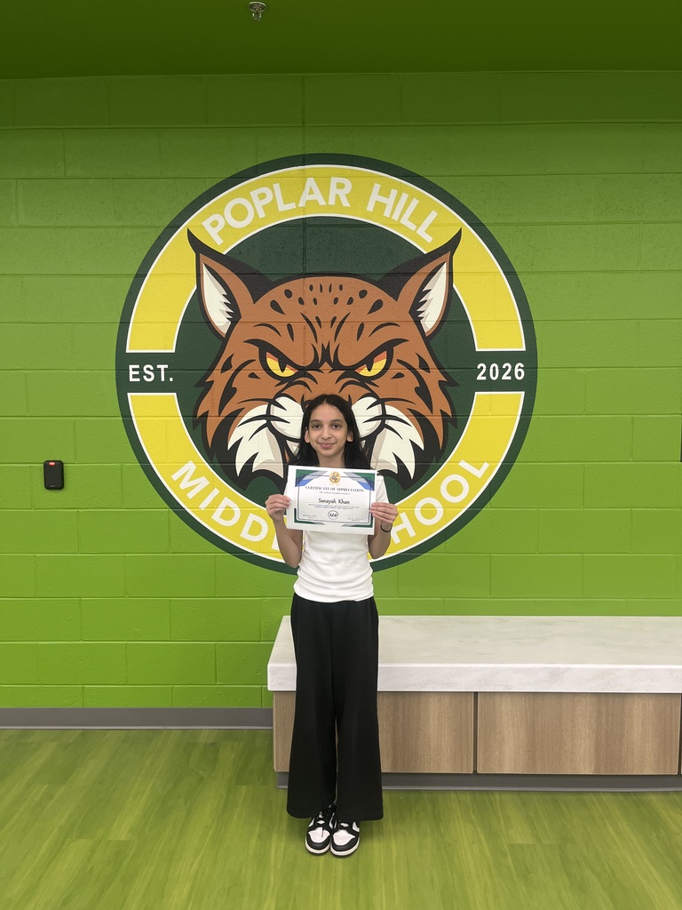 A girl in a white shirt and black pants stands before a green wall displaying the Poplar Hill Middle School emblem.