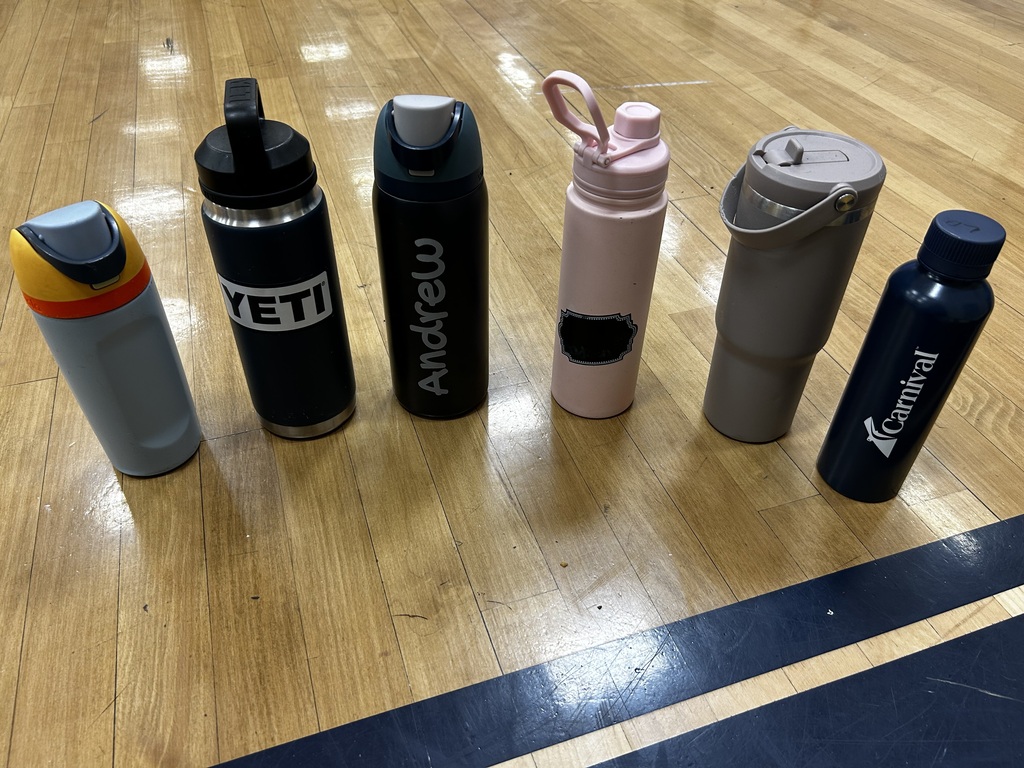 Seven water bottles in various colors and sizes are lined up on a wooden floor. The bottles have personal names or brand names.