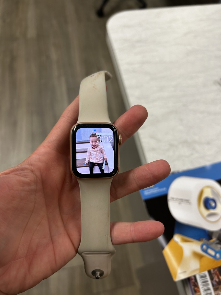 A hand holds a watch with a picture of a child on the screen. A white table and various items are in the background.