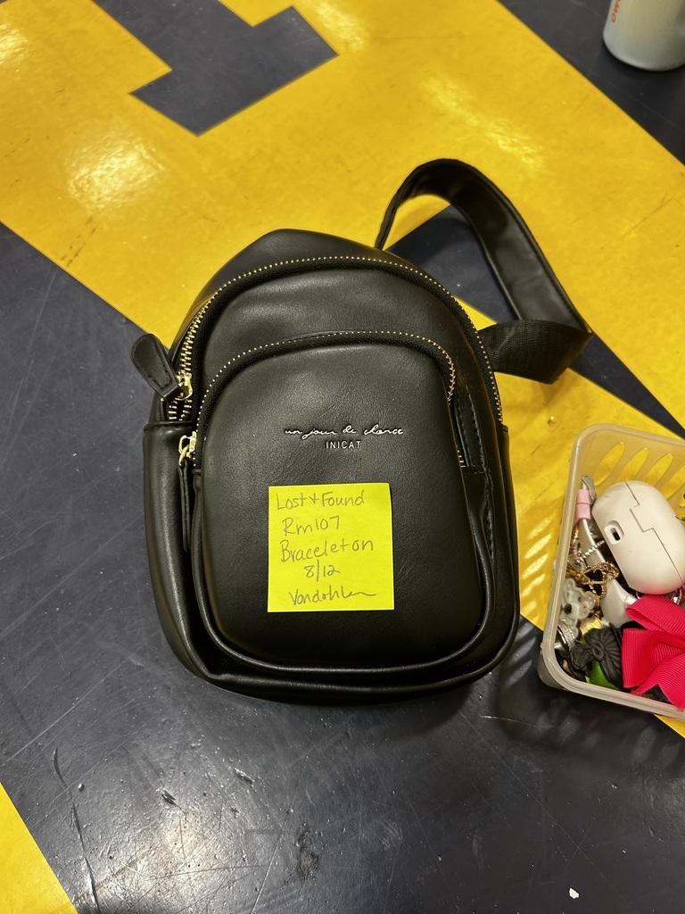 A black shoulder bag with a yellow note is on a yellow and black floor next to a clear container holding various small items.