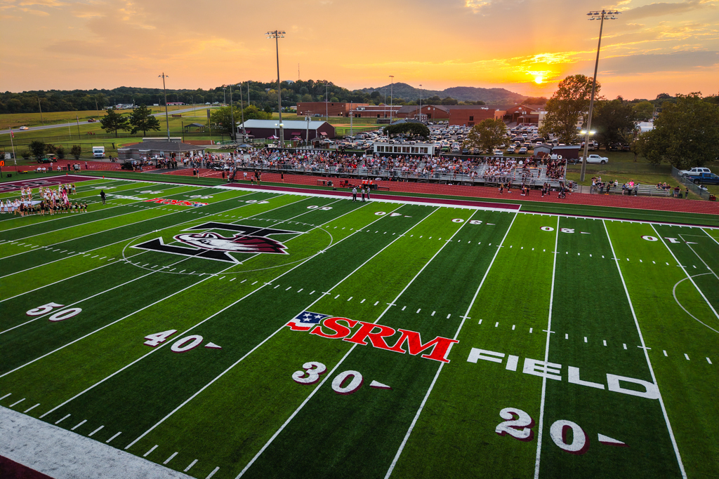 A football field with numbers and a logo, surrounded by stands, at sunset with a warm glow.