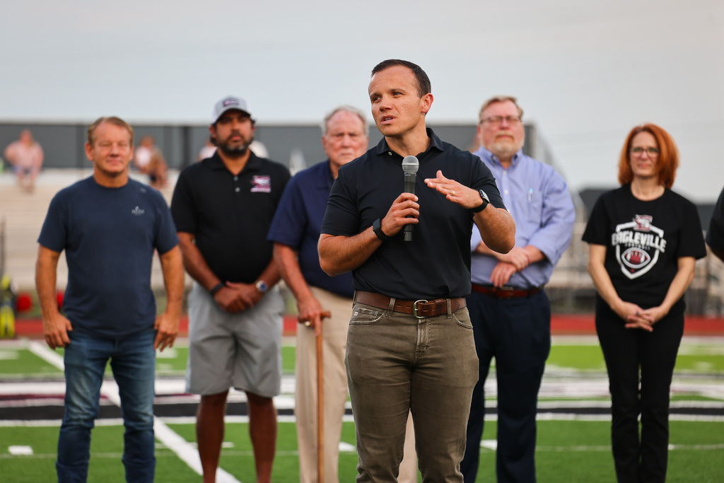 A man speaks at a microphone on a football field, surrounded by six others. They are all wearing T-shirts.
