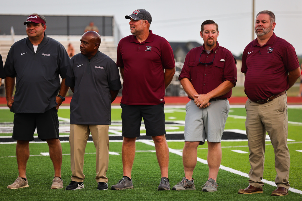 Five men stand on a football field, all wearing shirts and shorts, with one holding a camera.