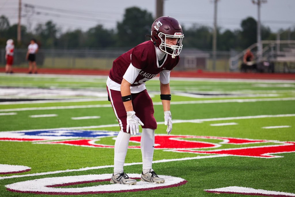 A football player in maroon and white gear stands on the field, helmet on, awaiting play.