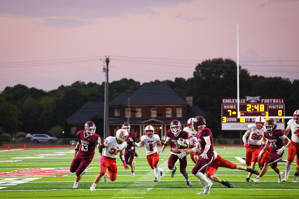 Football players wearing helmets and jerseys run across a field with a scoreboard in the background.