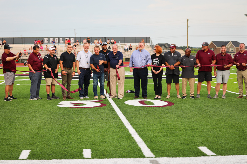 A group of people on a football field holds a ribbon. Some wear hats, sneakers, and casual attire.