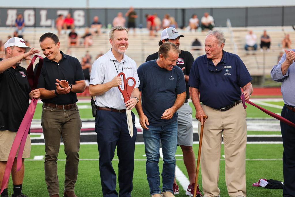 Six men stand on a football field holding ribbons. One uses scissors to cut a ribbon. Behind them, people sit on the bleachers.