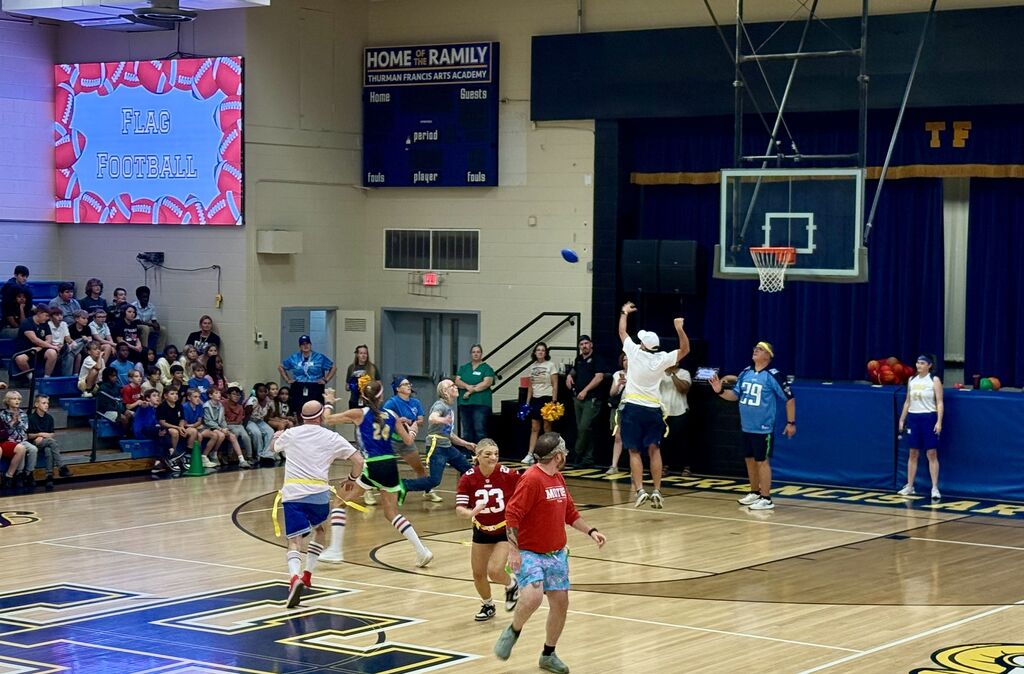 Players in shorts and jerseys play a game on a basketball court with a scoreboard and a cheering crowd.