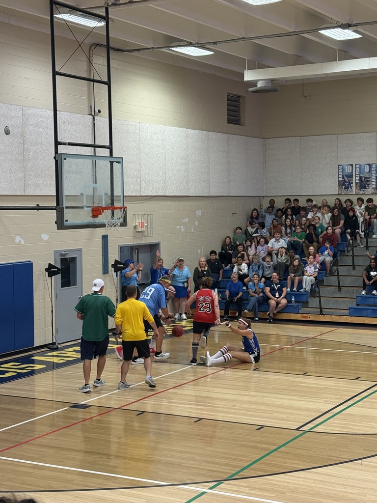 A basketball court with players, one dribbling, others watching, in a gym with blue bleachers.