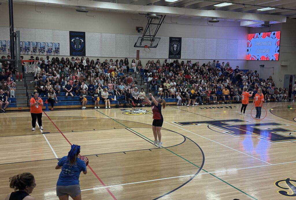 A basketball court with players in colored jerseys. One player holds a ball. The audience is seated on the bleachers.