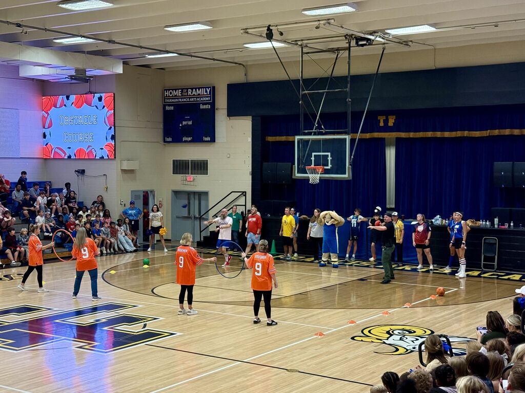 An indoor basketball court with players in orange jerseys. Blue walls, large screen, audience seated.