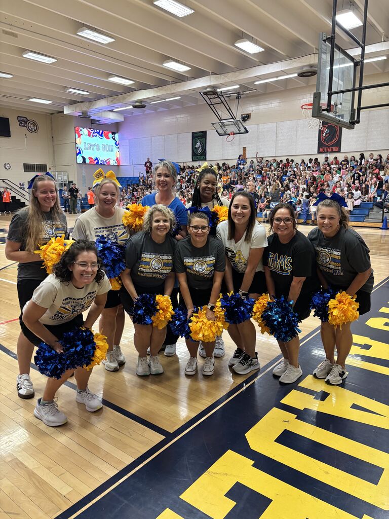 Cheerleaders pose on a basketball court with yellow and blue pom-poms. Audience in the background.