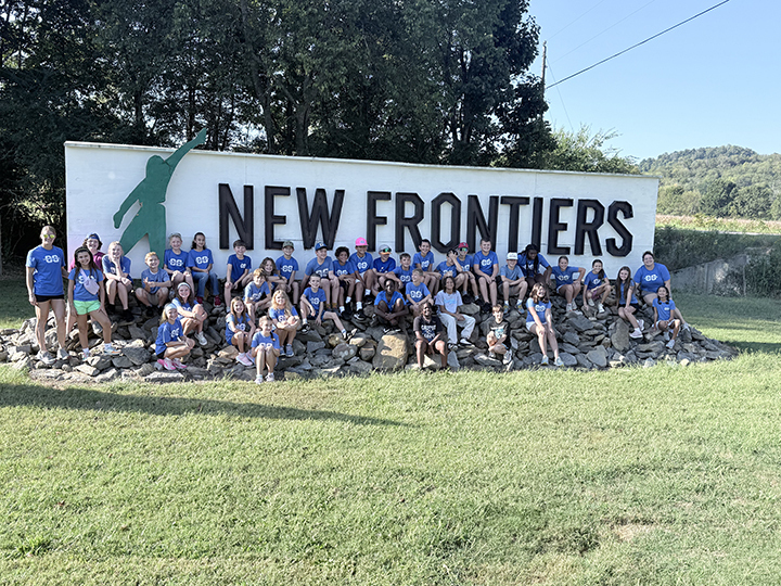 Group of children in blue shirts posing in front of a large "New Frontiers" sign.