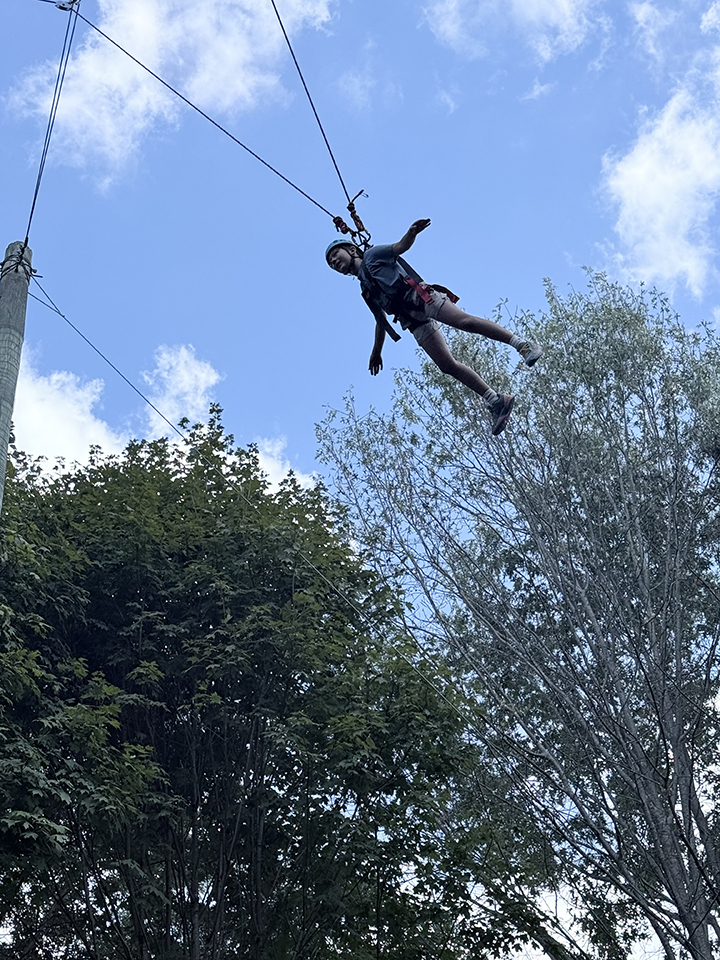 Person hanging from a zipline with trees and blue sky background.