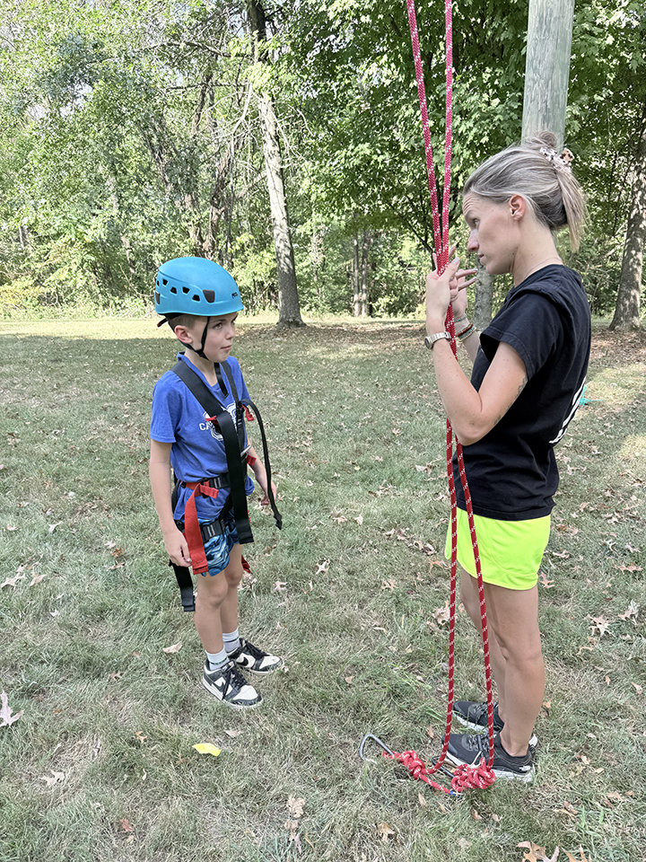 A child wearing a blue helmet, harness, and sneakers stands on grass, while a woman helps them with a red rope.