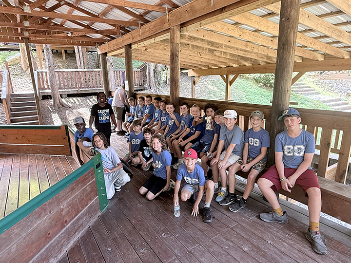 Group of young people in blue shirts and caps sit and stand on a wooden porch with a metal roof.