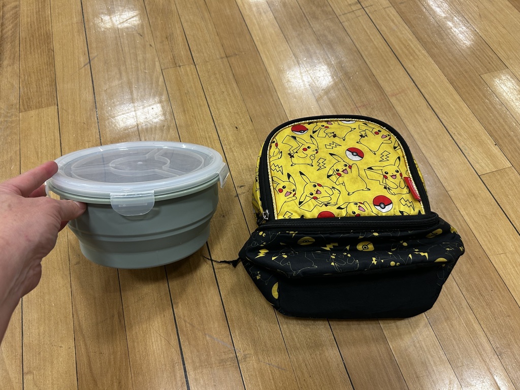 A person holds a grey container with a lid next to a black and yellow backpack with a Pikachu design. Both are on a wooden floor.