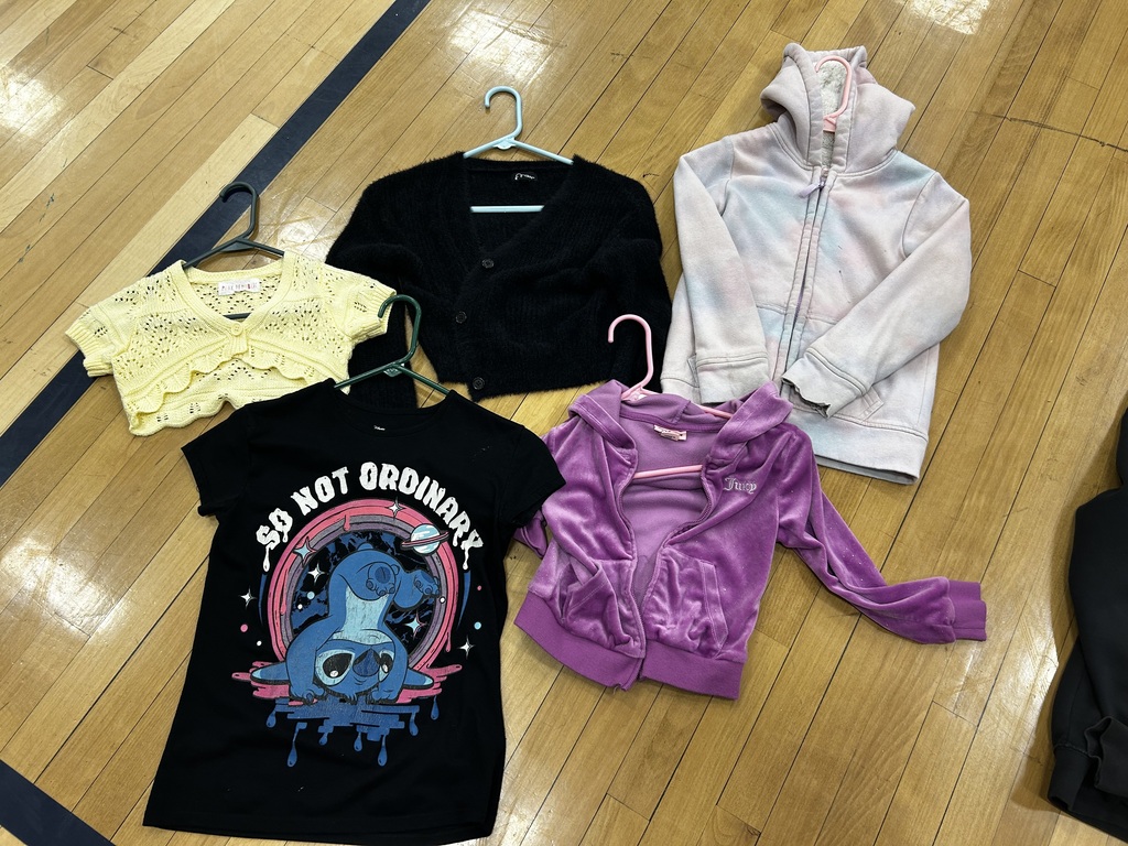 Five tops, including a black graphic tee, yellow lace top, black cardigan, pink hoodie, and white hoodie, lie on a wooden floor.