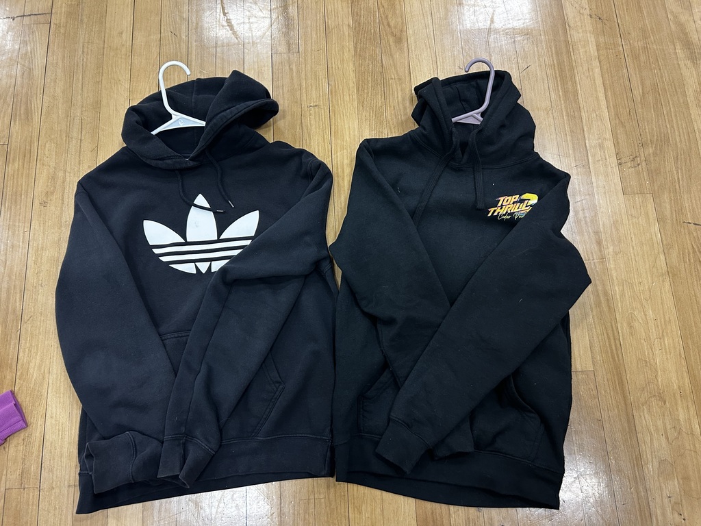 Two black hoodies on hangers on a wooden floor, one with a white Adidas logo and the other with yellow text.
