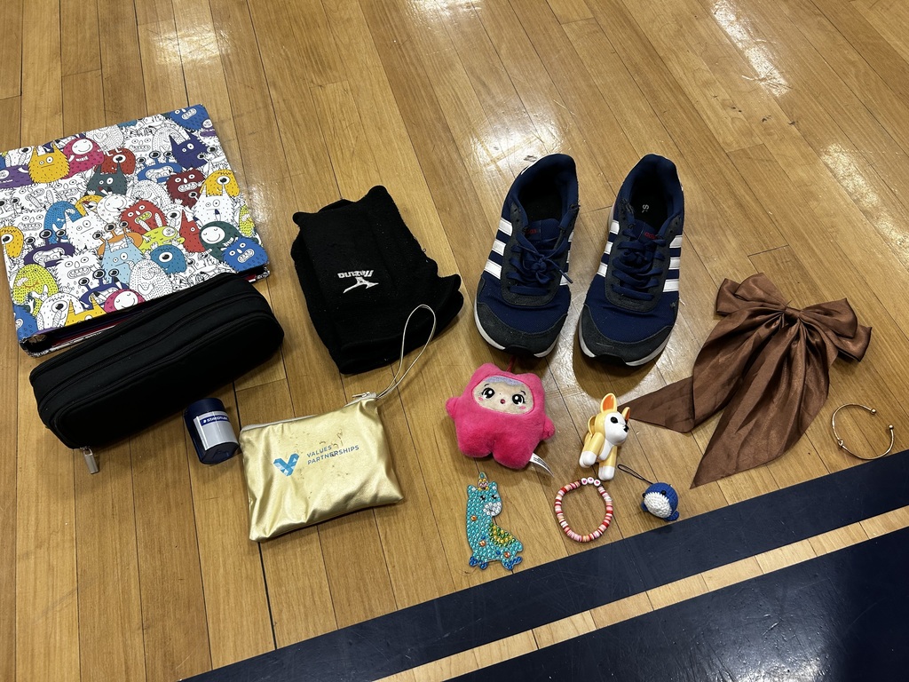 Various items on a wooden floor include a pencil case, a pouch, shoes, a bow, a bottle, a stuffed animal, and a small toy.