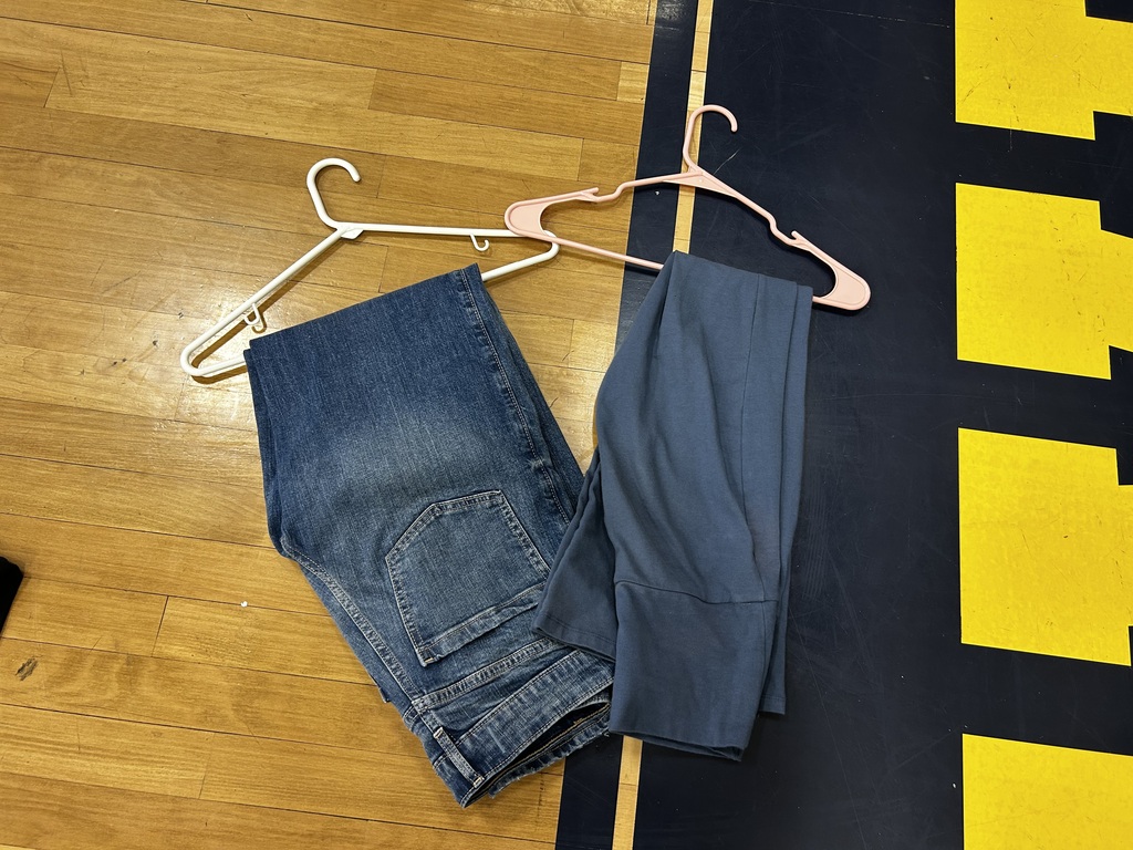 Two pairs of pants, one denim and one gray, on a white hanger against a wooden floor with a black mat.