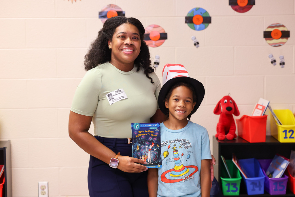 A woman and a young boy stand in a classroom with colorful decorations and shelves with bins.