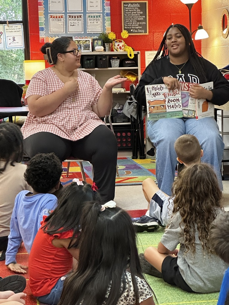 Two women sit in chairs with children in front of them, reading a book. A rug and shelves are visible in the background.