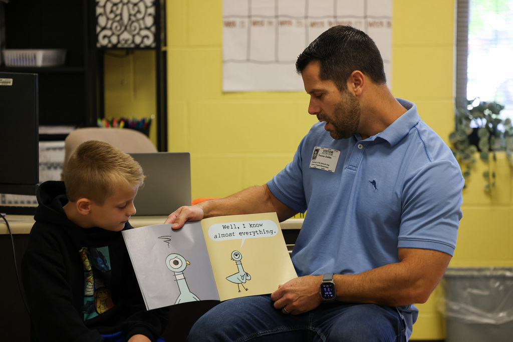 A man in a blue shirt reading a book to a boy in a black hoodie.