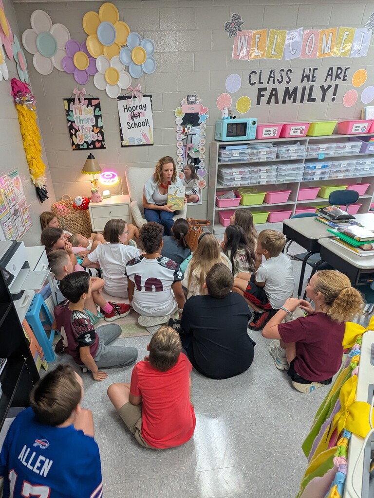 Classroom with students sitting on the floor listening to a teacher read a book. Colorful decorations, shelves, and chairs are present.