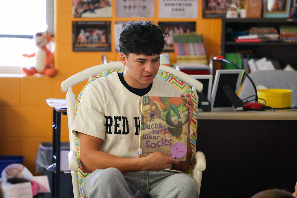 Person in a white shirt reading a book. Background includes classroom walls, desk, books, and a toy.