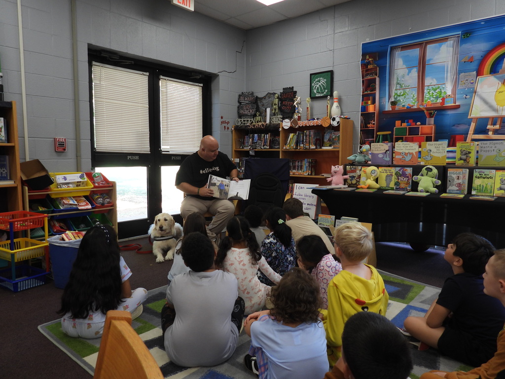 A man reads to a group of children in a classroom. Bookshelves are behind them, and a dog sits nearby.