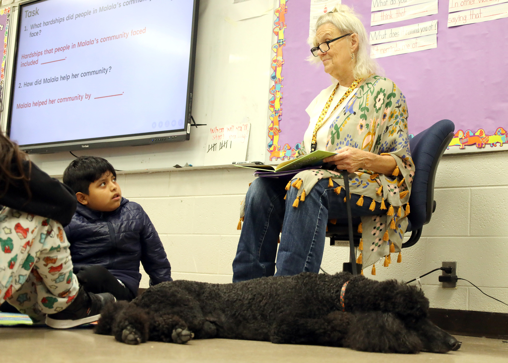 Woman seated on a chair, reading to a boy and girl seated on the floor. A dog lies at their feet.
