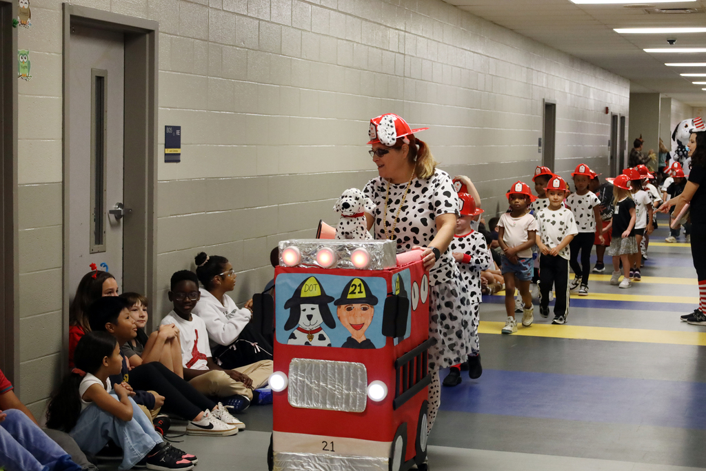 A person in a costume and a fire engine cart walks down a school corridor, followed by children.