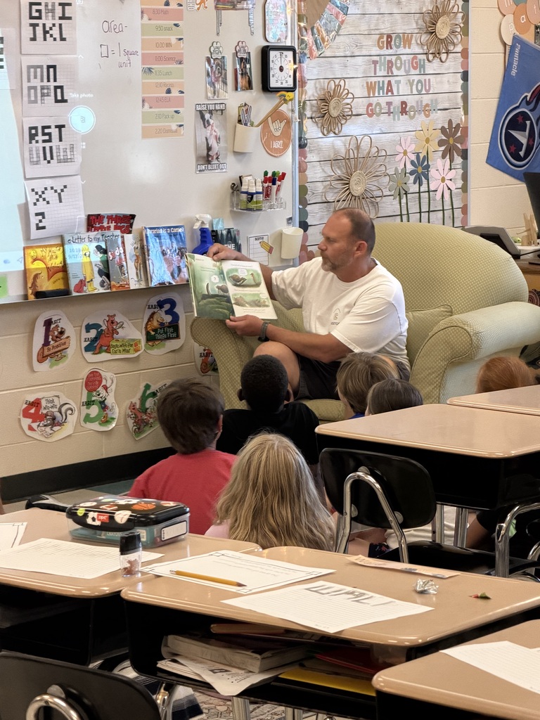 A classroom with a teacher reading a book to children seated in chairs. Desks are arranged in rows.