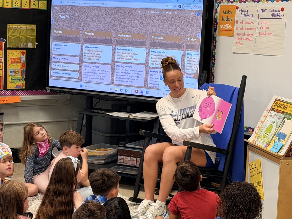 Woman sits on blue chair in front of class with students sitting on floor. Woman reads from pink book.