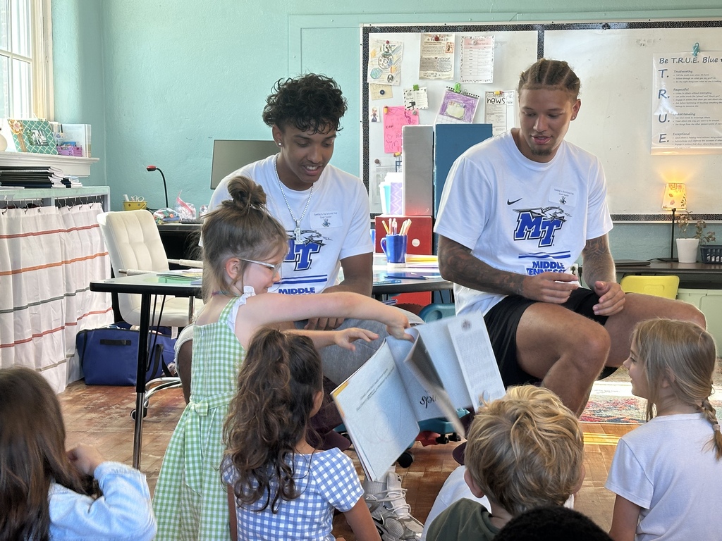 Two adults in matching shirts sit cross-legged on a rug with children. One adult reads a book to the children.