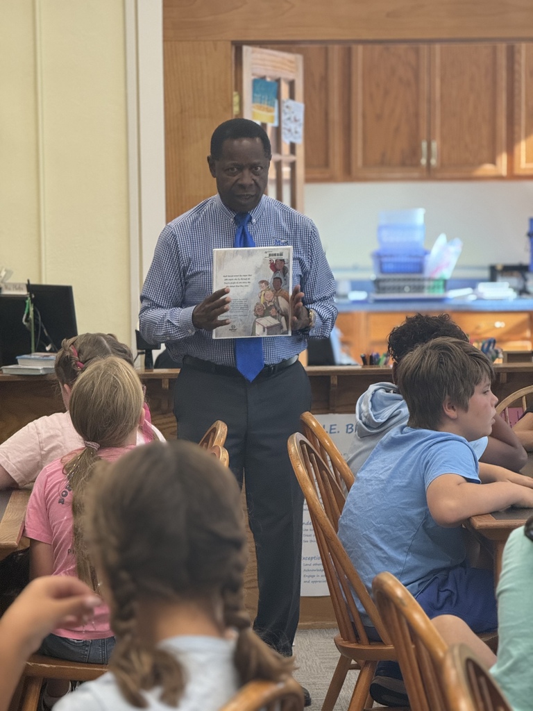 A man in a blue shirt and tie reads a book in front of a classroom.