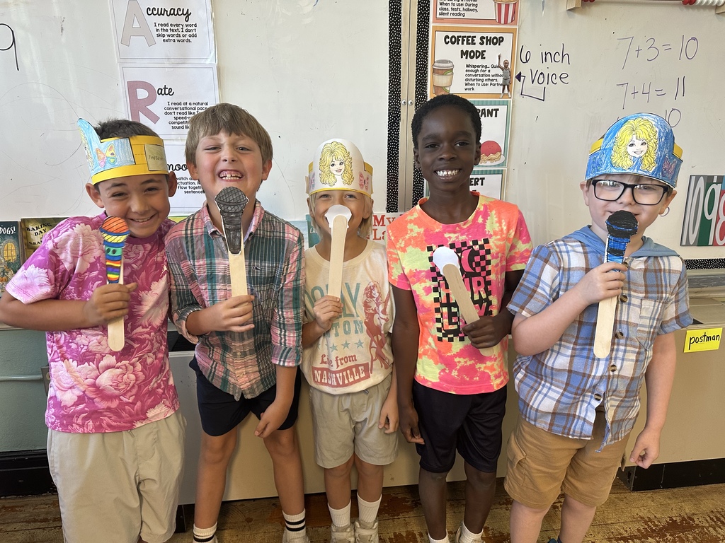 Children dressed in hats and holding microphones stand in a classroom with a whiteboard behind them.