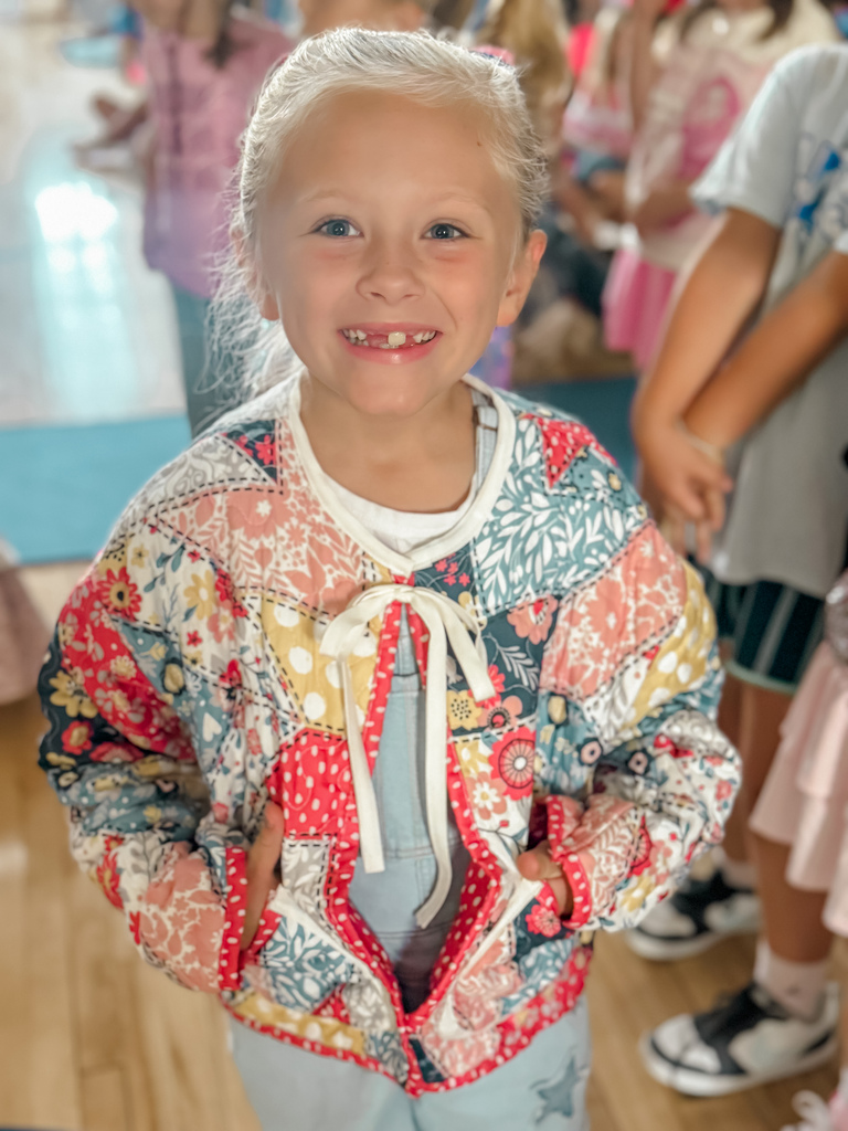 A child with blond hair smiles, showing a gap in their teeth. They wear a colorful patchwork jacket over a white shirt and blue pants.