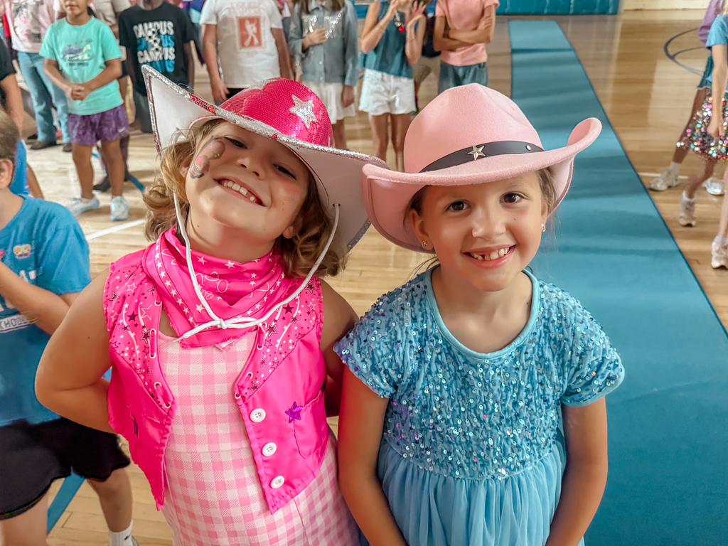 Two young girls in pink cowboy hats stand next to each other in a gym. Other children stand behind them.