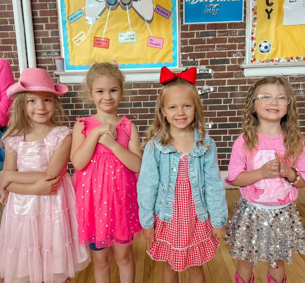 Four young girls, two in pink dresses, one in a denim jacket, and one in a glittery skirt, stand smiling and making hand gestures.