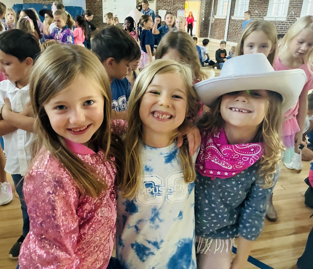 Three girls in a school gymnasium. The girl in the center wears a cowboy hat and scarf. The girl on the right wears a pink shirt.