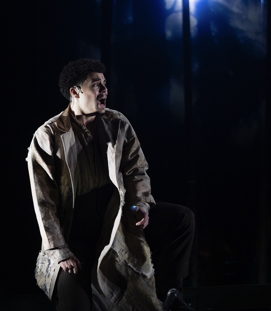 A man, wearing a trench coat, is kneeling on a stage, mouth open. Spotlight shines from above.