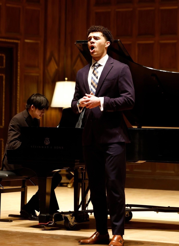 A man in a suit and tie is singing in front of a piano, with another person playing.