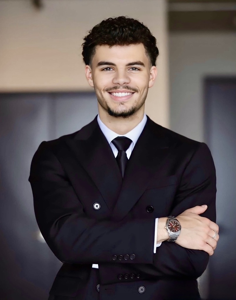 Person in a black suit and tie, smiling, standing with arms crossed, possibly in a professional setting.